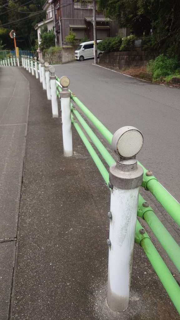 白いポールと緑色の柵が手前から奥へ続く住宅街の道路
